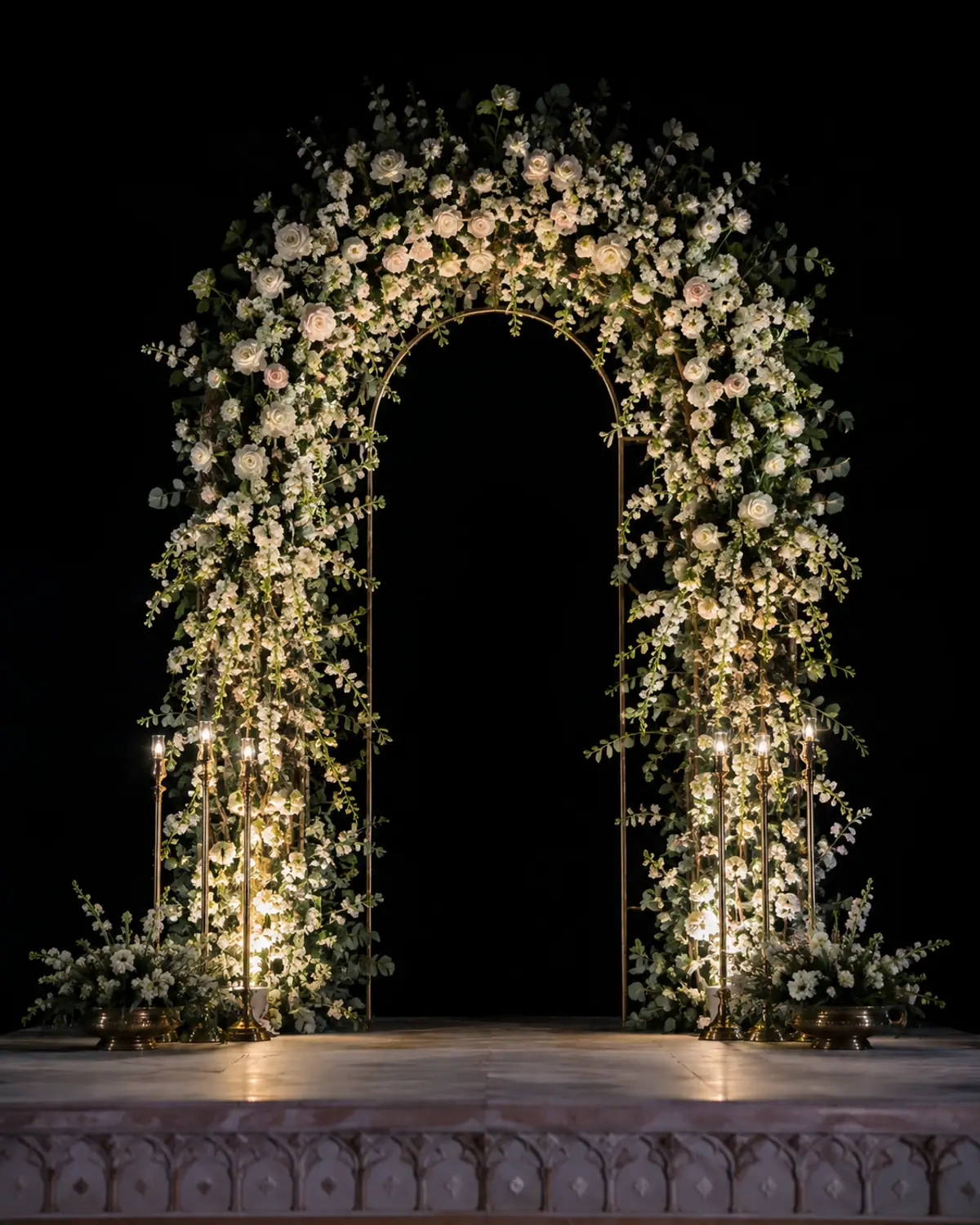 A tall floral arch of white and blush roses, uplit by brass candle stands against an unlit backdrop.